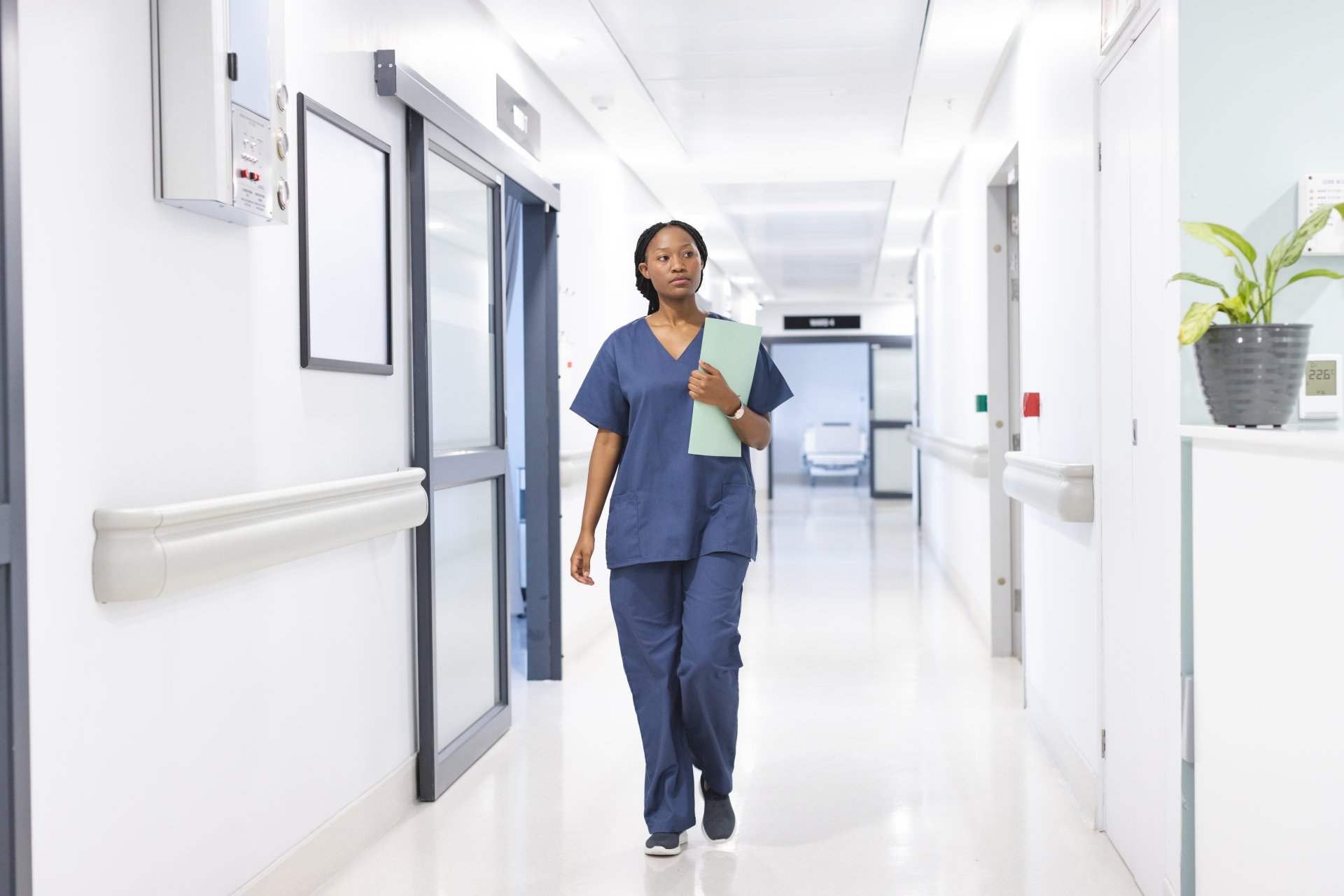 A young female healthcare worker walks down an empty hospital corridor holding documents.