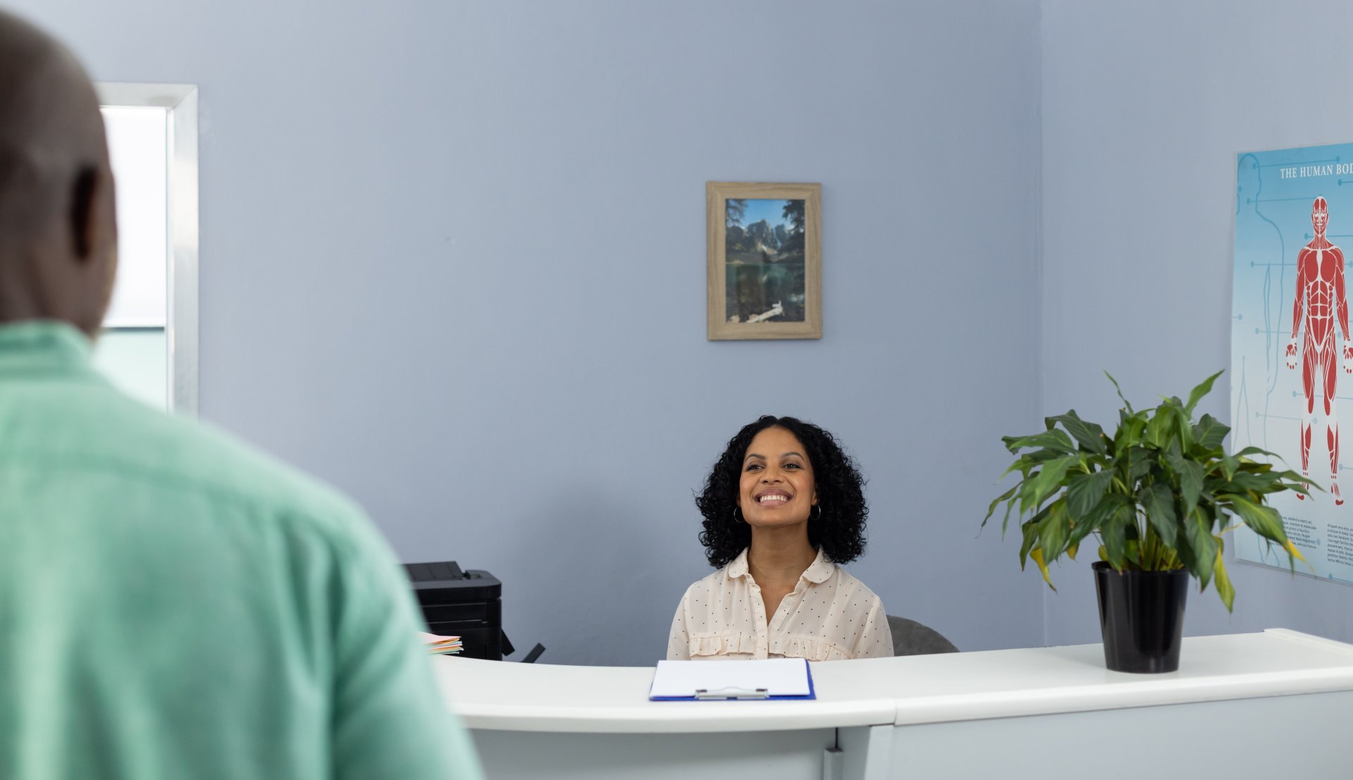 A woman behind the front desk of a healthcare professional's office smiles at a male patient approaching her.