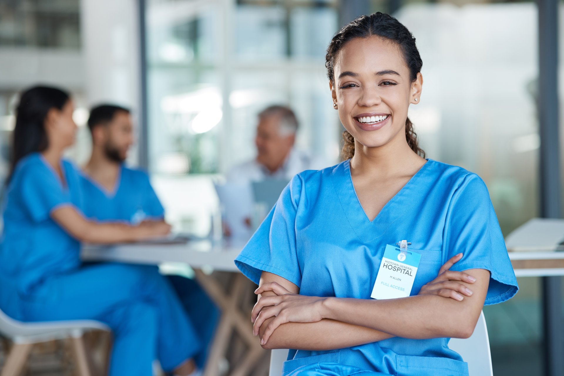 A young female healthcare worker poses and smiles while her coworkers hold a group discussion behind her.