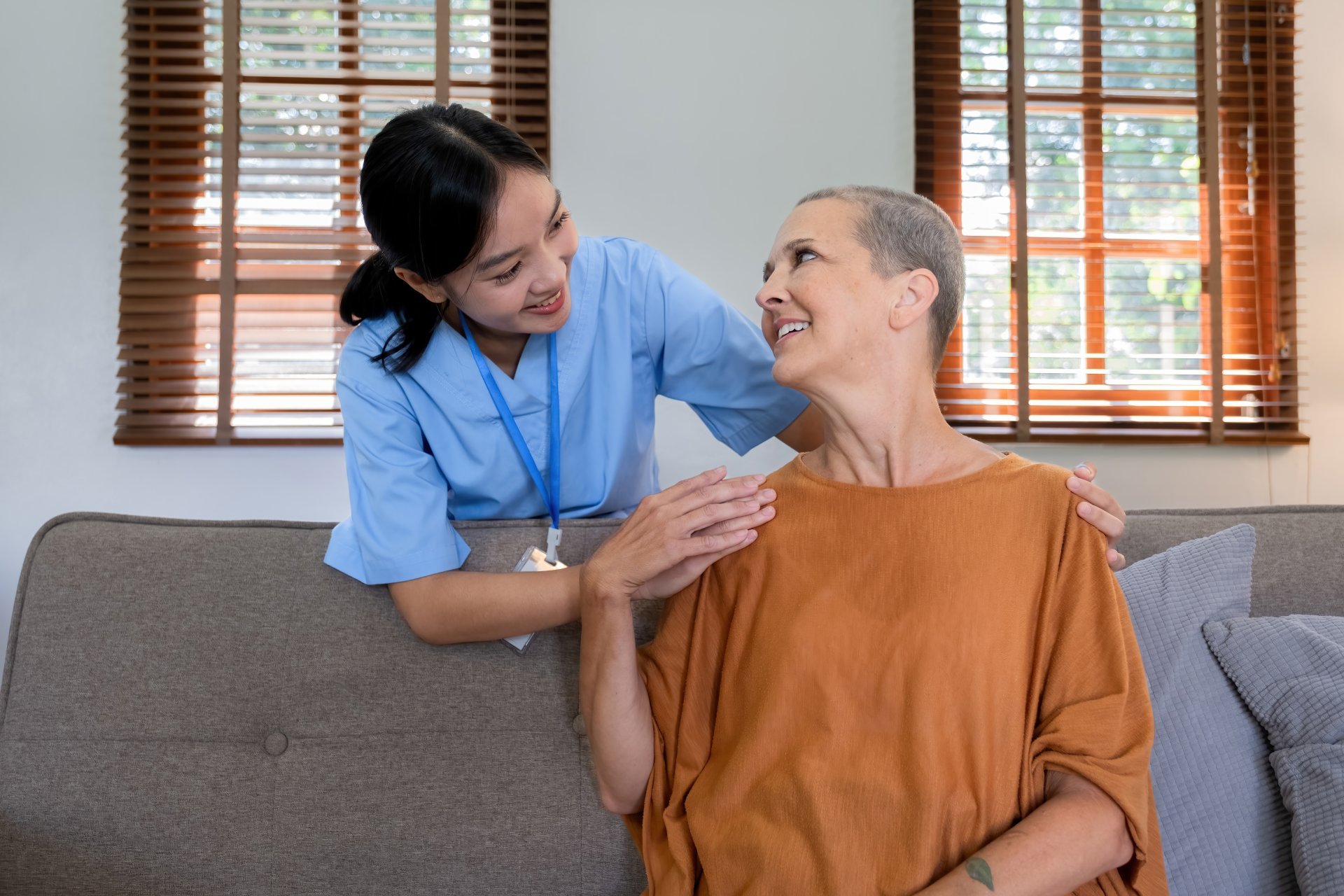 A young caregiver in scrubs and a lanyard checks on her elderly female client in her home.