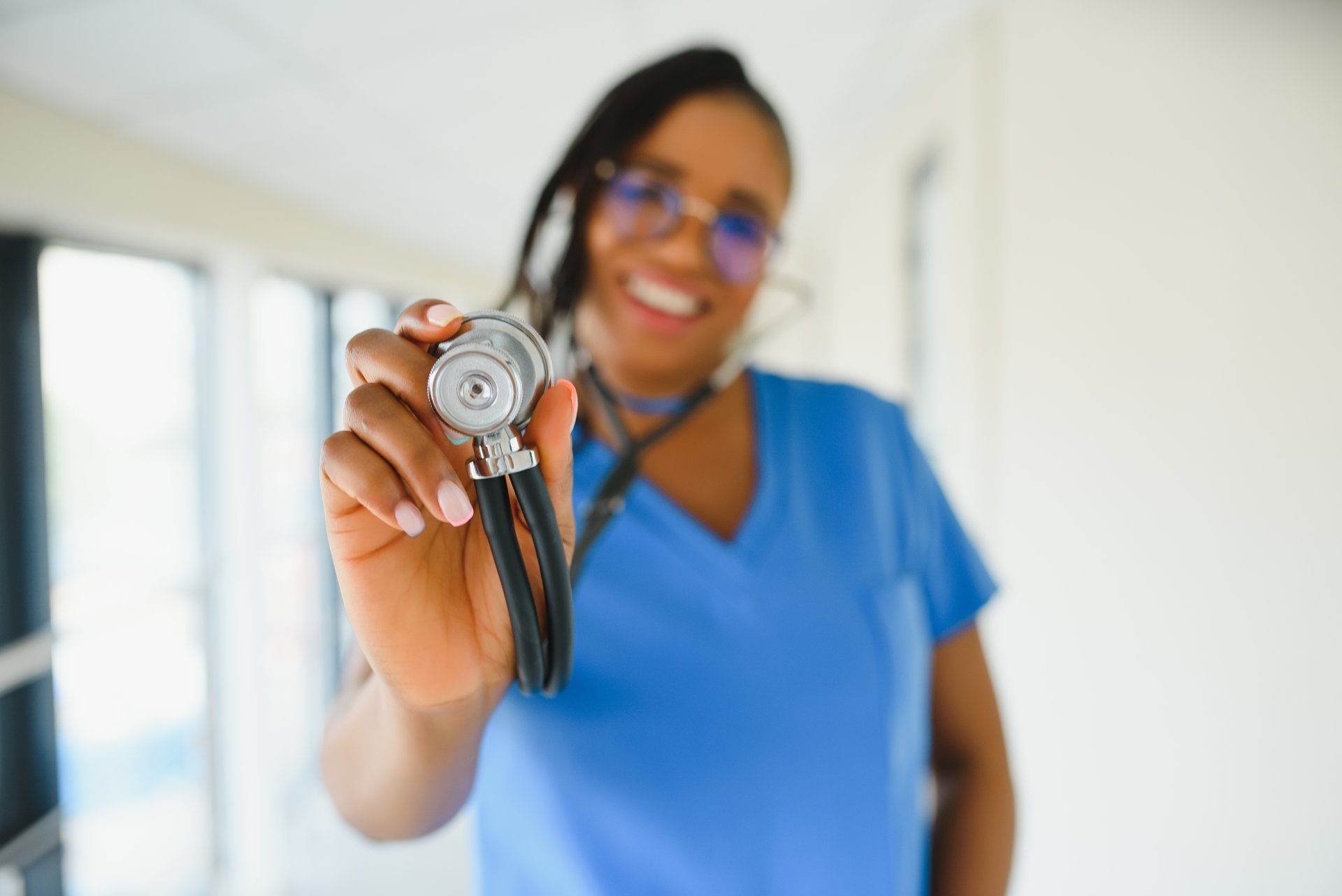 A female nurse holds her stethoscope up to the camera and smiles.