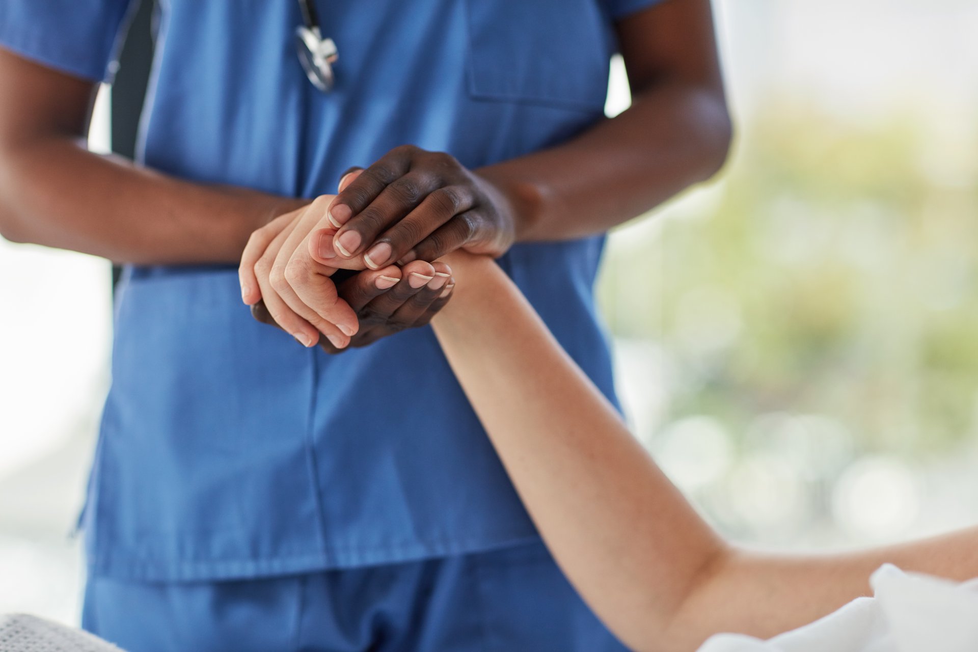 A clinician wearing scrubs and a stethoscope holds their patient's right hand with both hands while standing at their bedside.