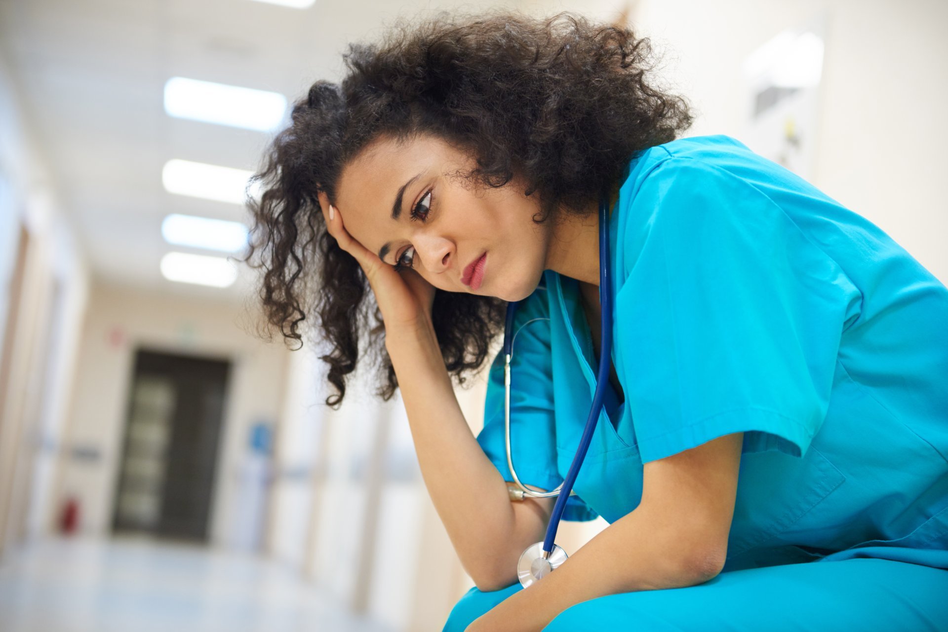 A young female clinician puts her hand to her forehead while sitting in a hospital hallway.