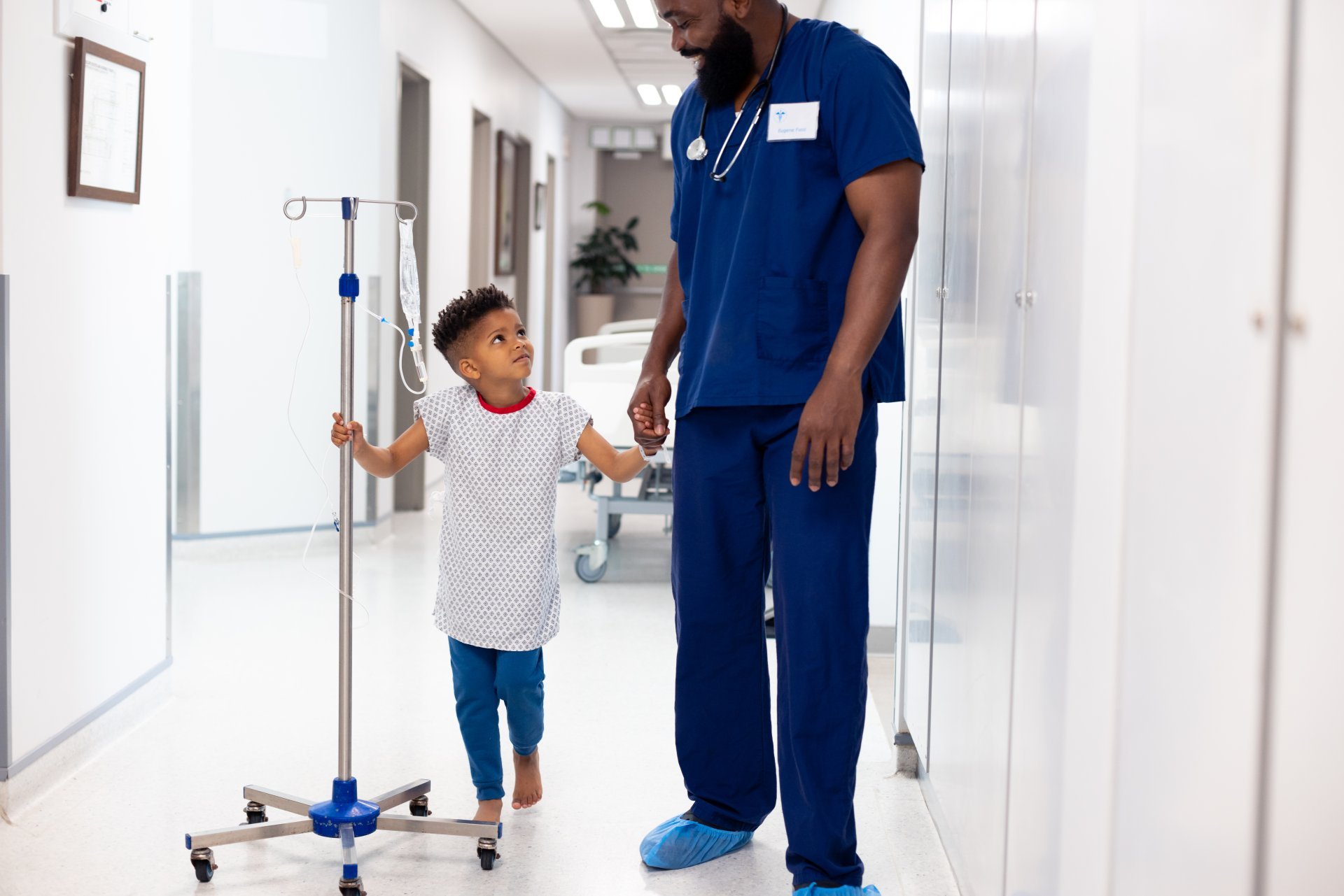 A young male nurse leads a small boy with an IV drip down the hallway of a hospital.