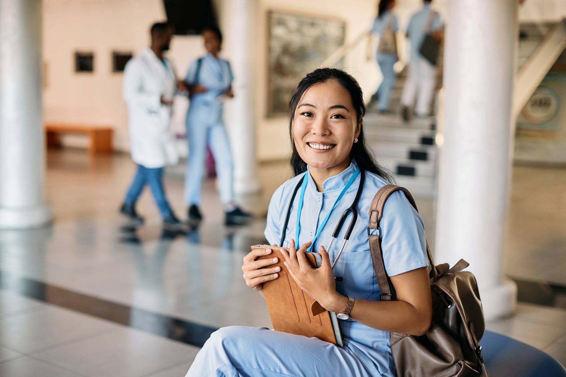 A young female nursing student smiles while sitting on a bench in the lobby of her school building.