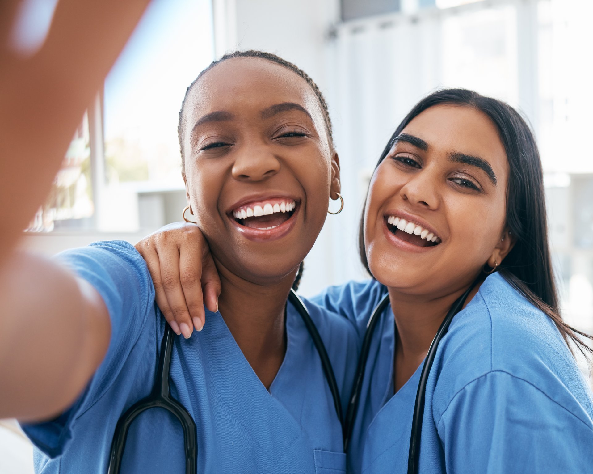 Two female nurses wearing scrubs and stethoscopes smile and pose for a photo together.