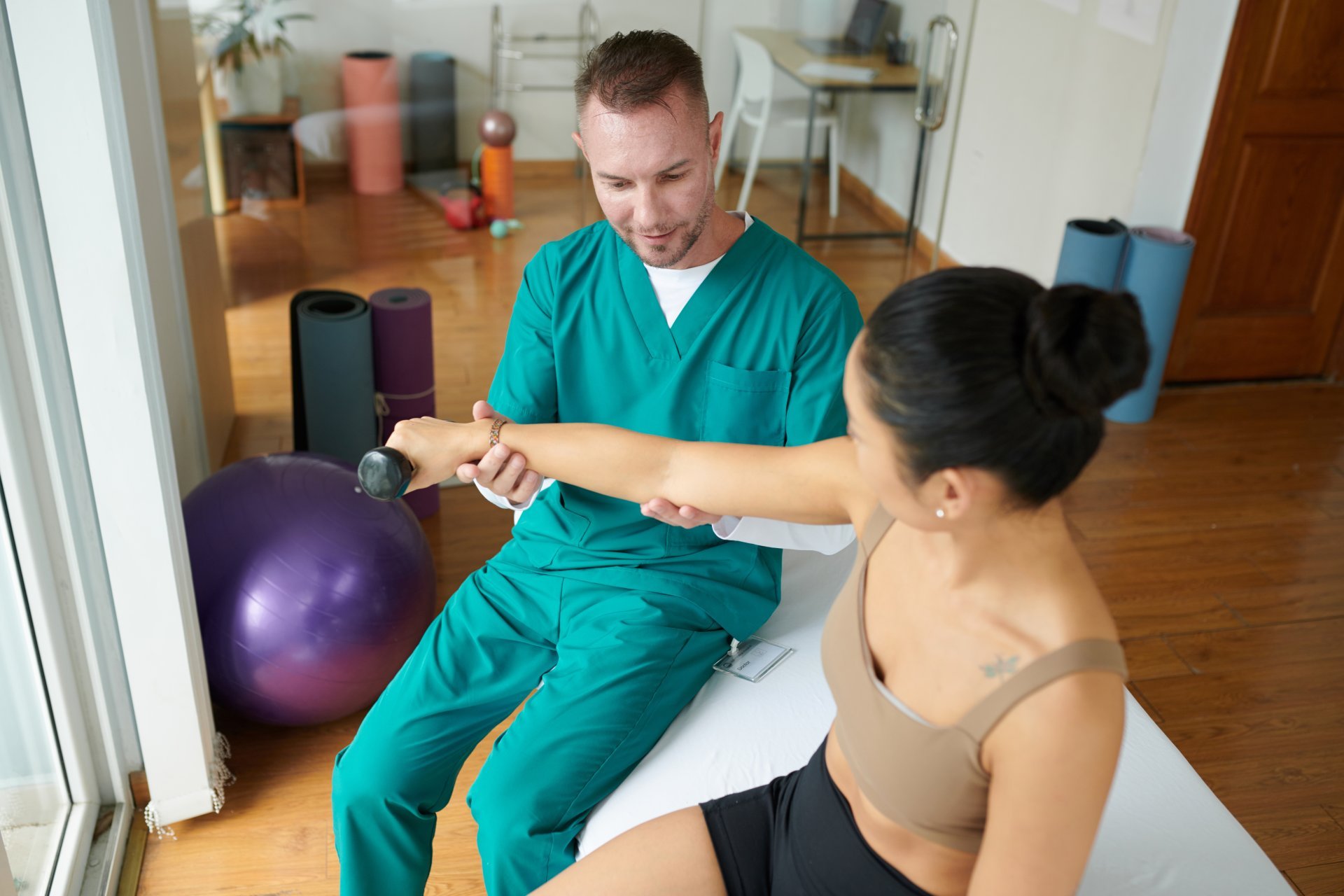 A young male physical therapist helps his young female patient rebuild arm strength using a dumbbell.