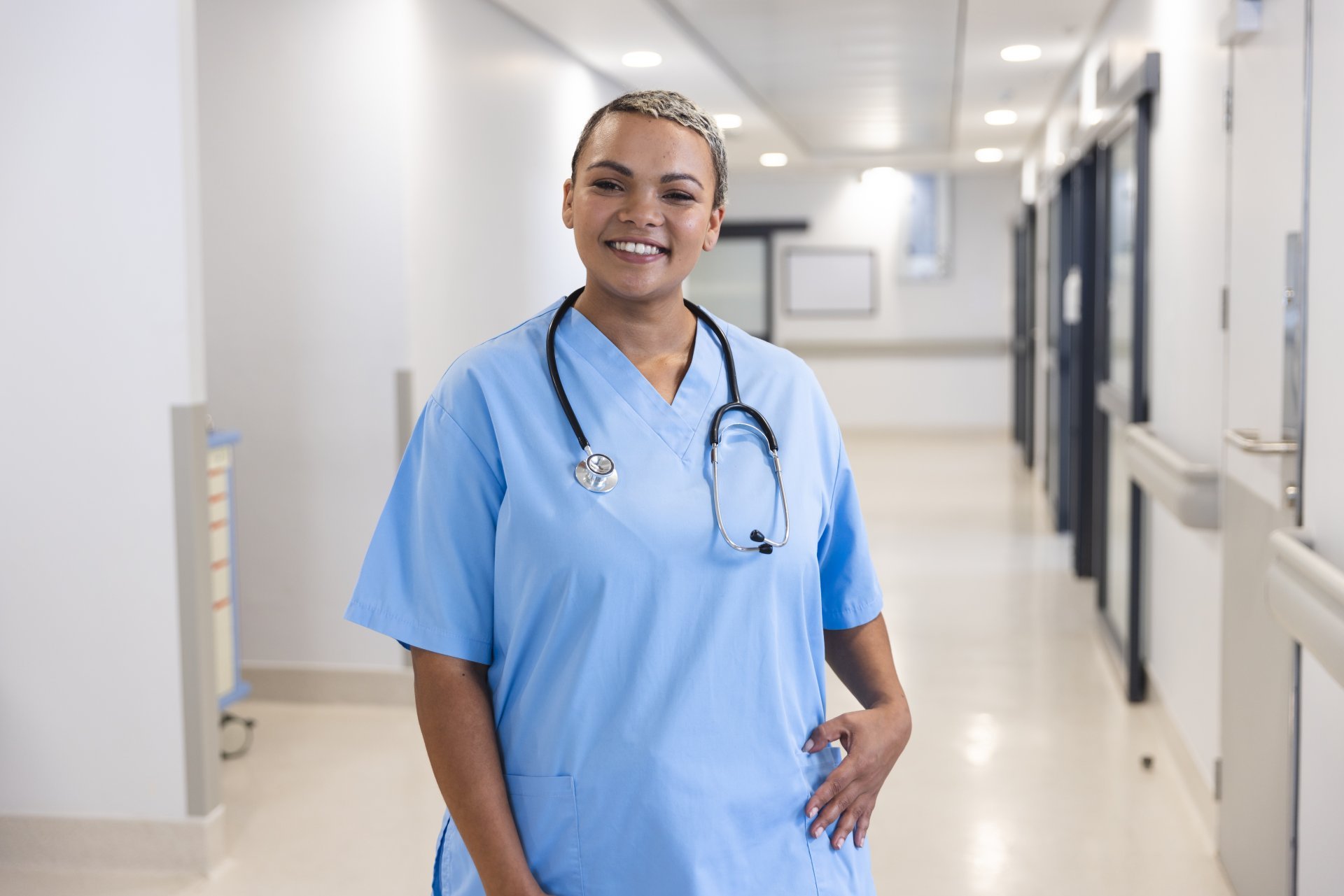 A smiling female nurse wearing a stethoscope stands in a hospital corridor. 