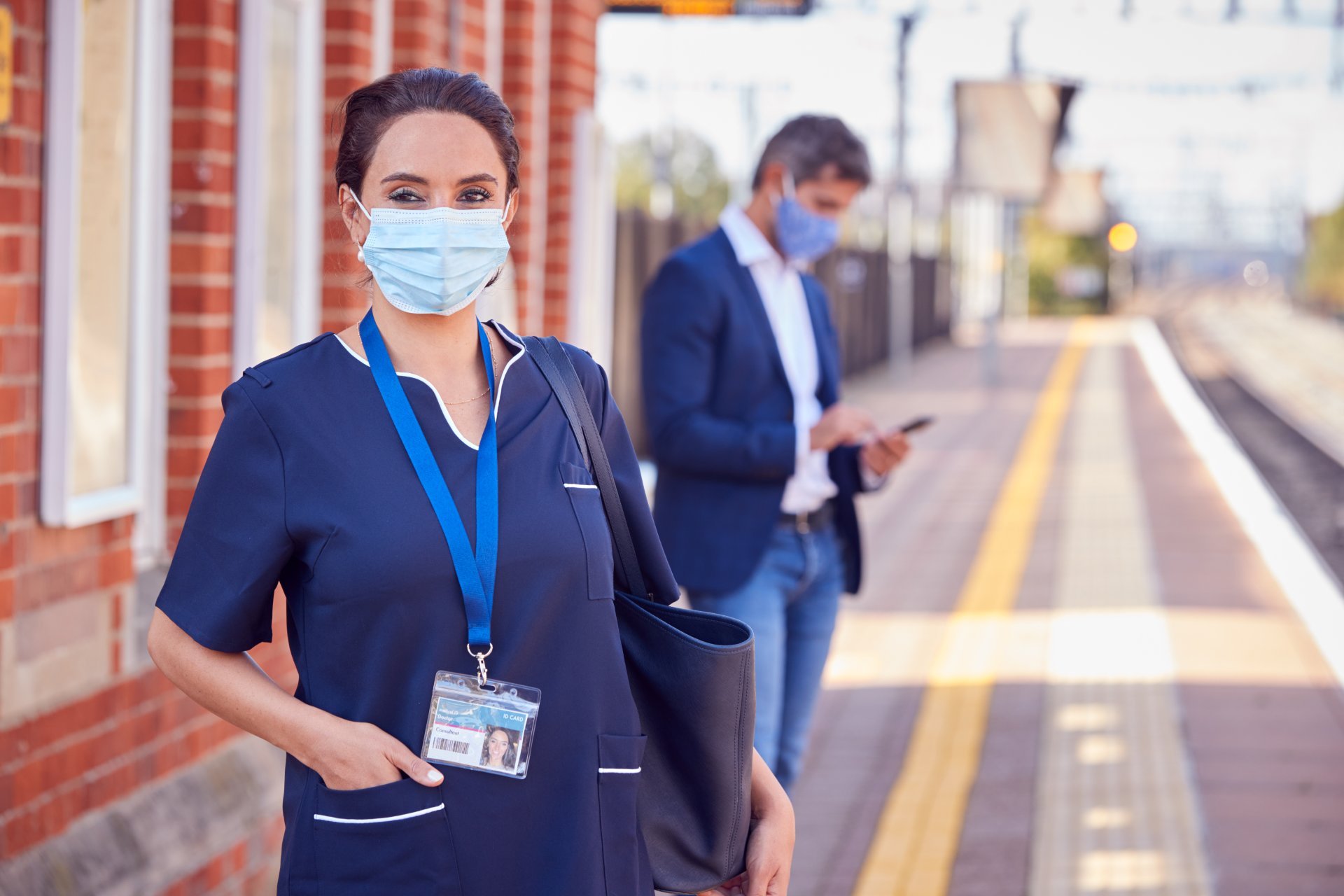 A nurse, wearing a mask, stands on a railway platform in front of an out-of-focus man in a mask and business casual attire.