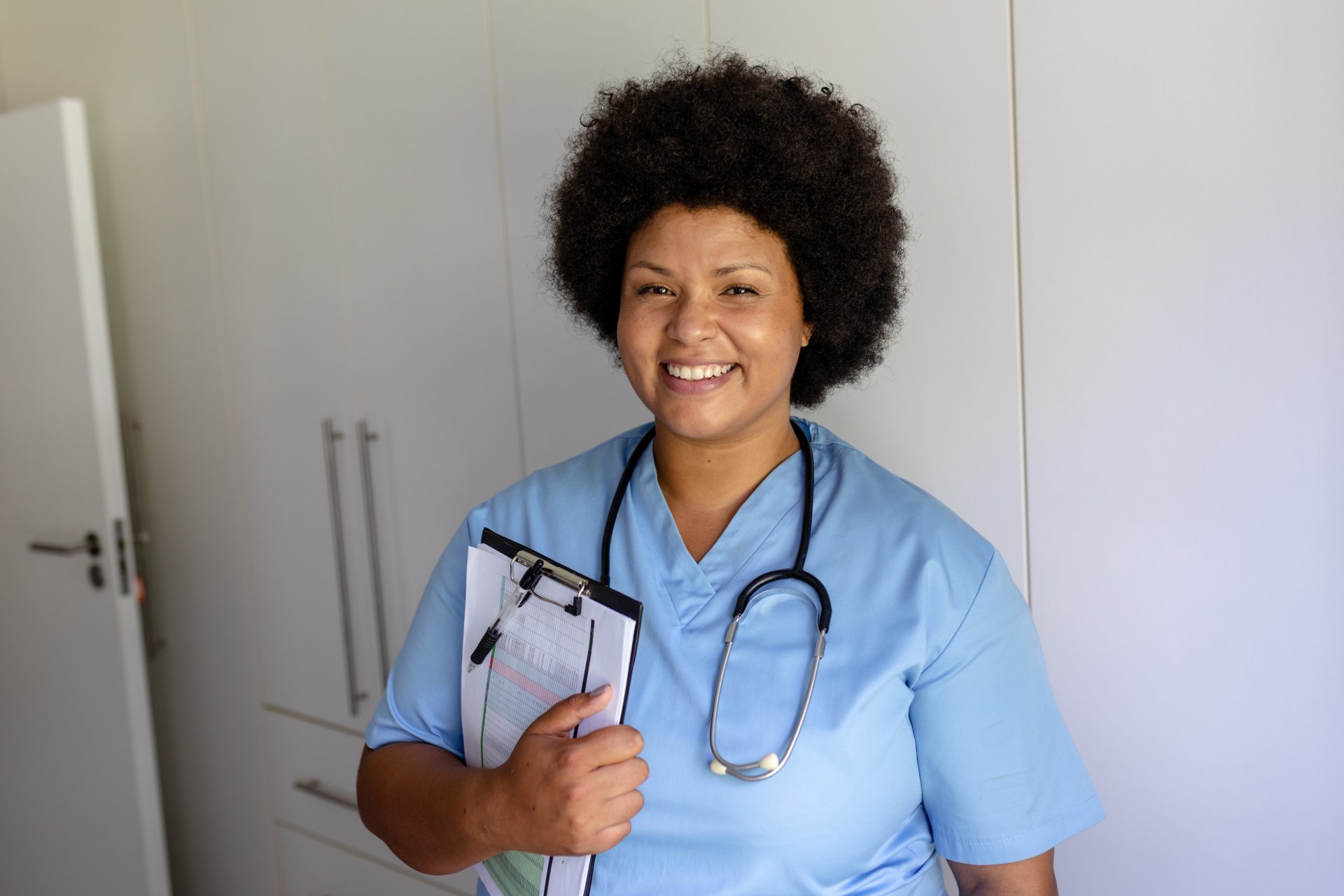 A smiling female clinician with a stethoscope holds a clipboard.
