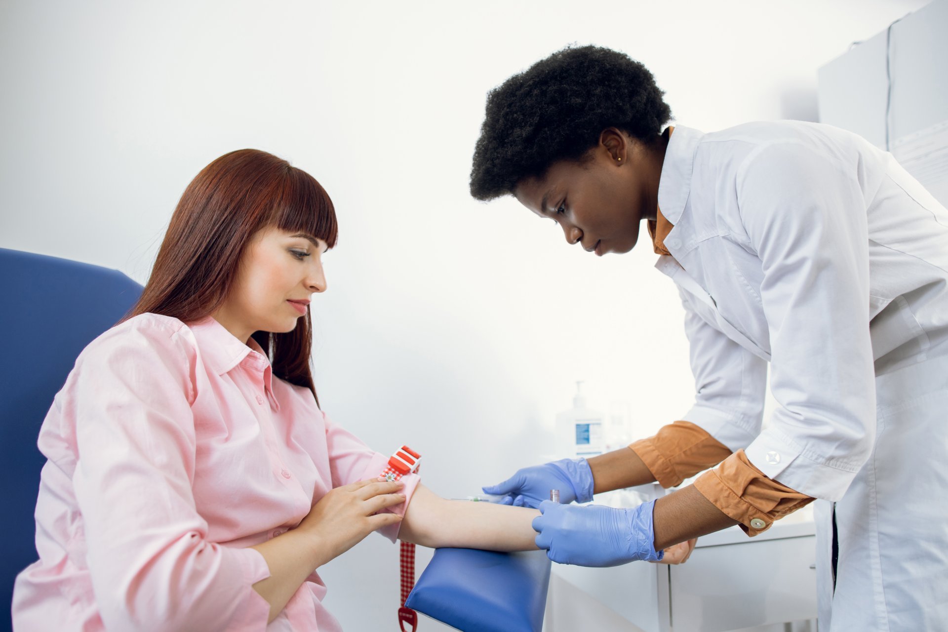 A female caregiver in a white lab coat and gloves prepares her female patient for a blood test.