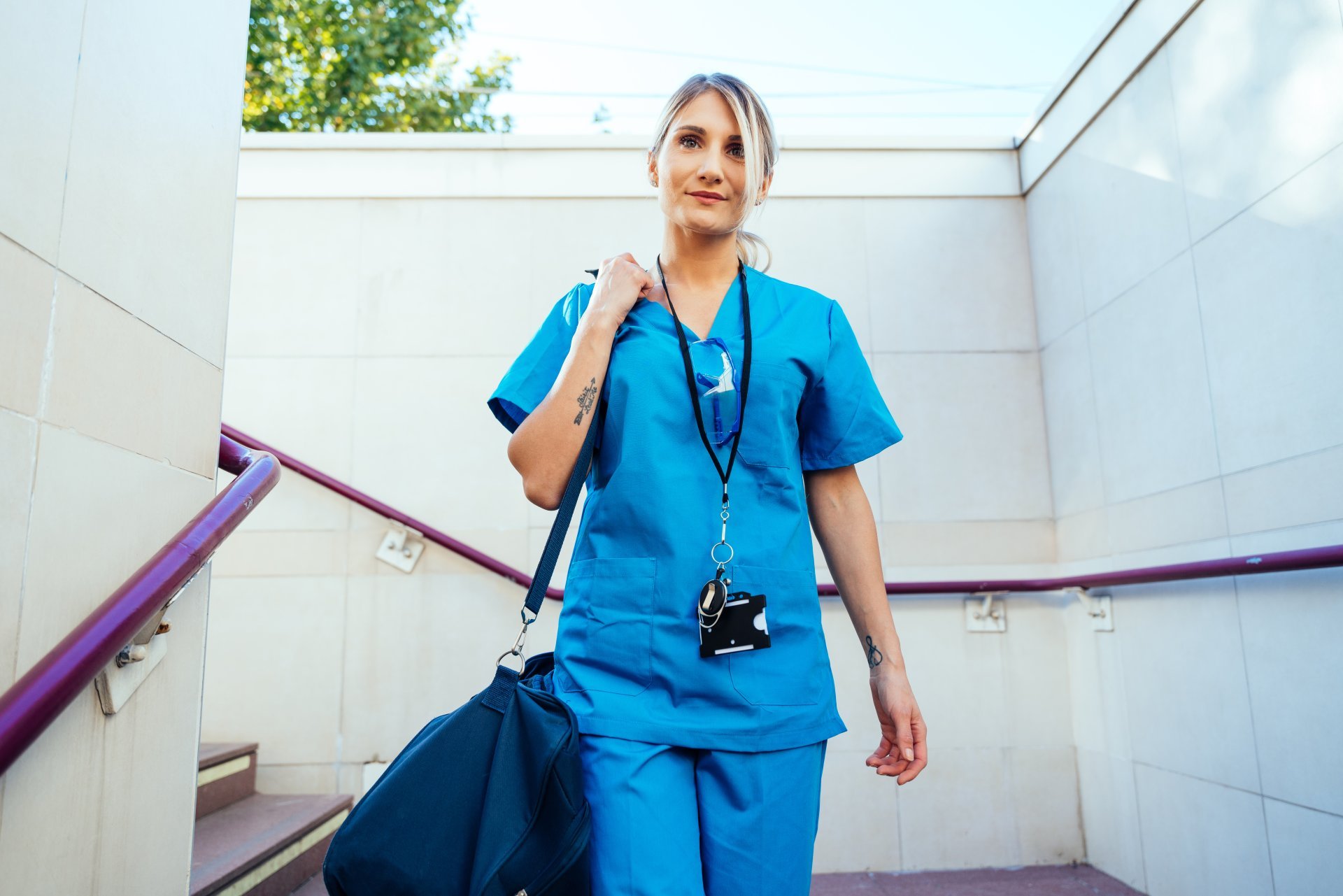 A female nurse in scrubs holds a duffel bag while descending an outdoor staircase.