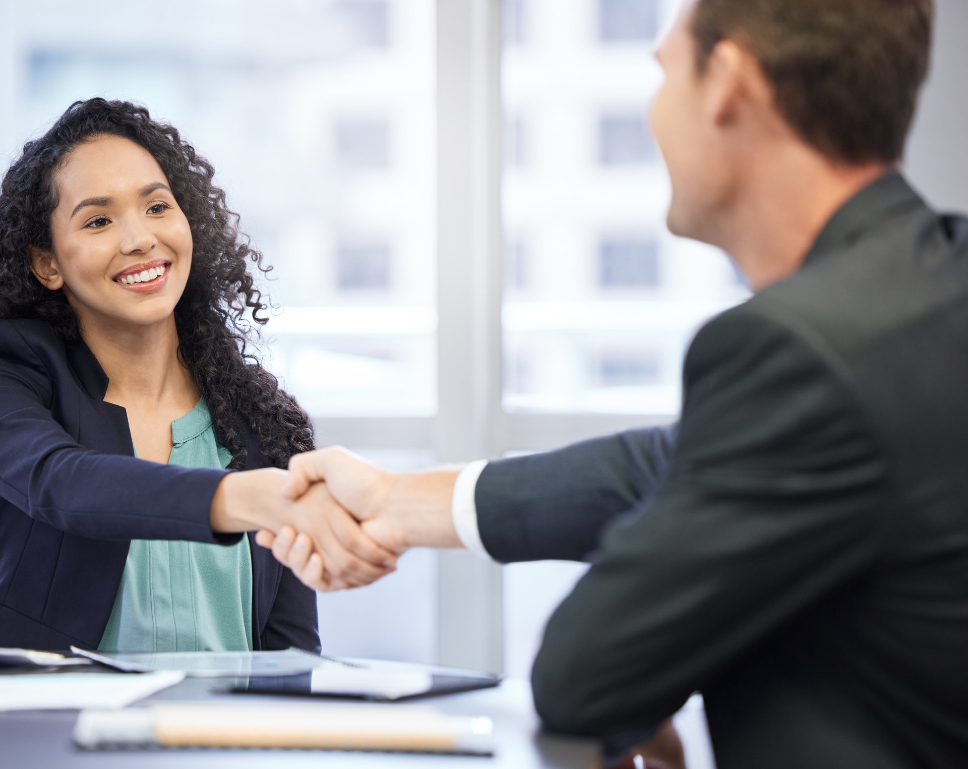 A young woman sitting behind a desk shakes the hand of an out-of-focus man in the foreground.