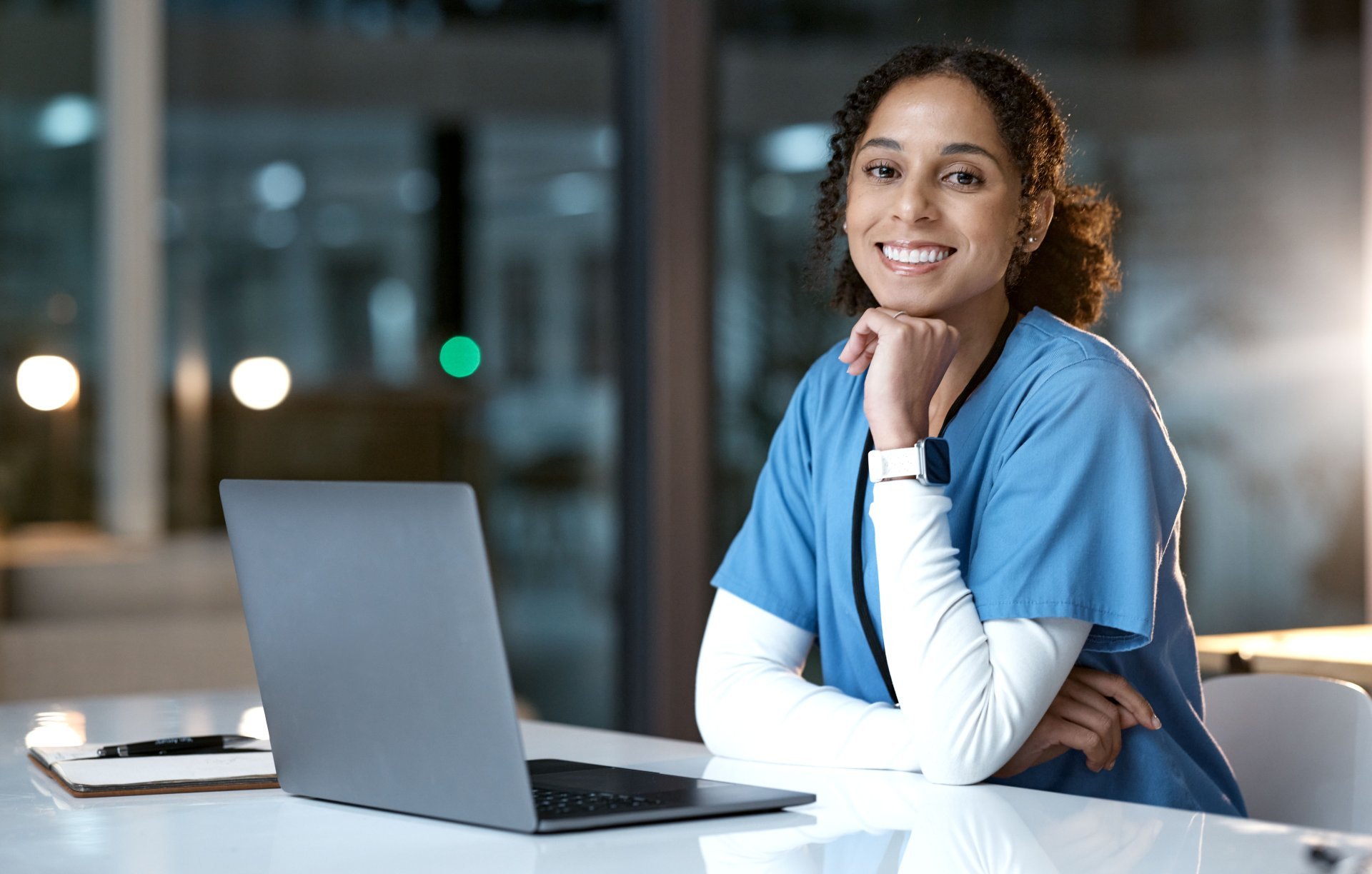 A smiling woman in scrubs sits at a desk in front of a laptop.